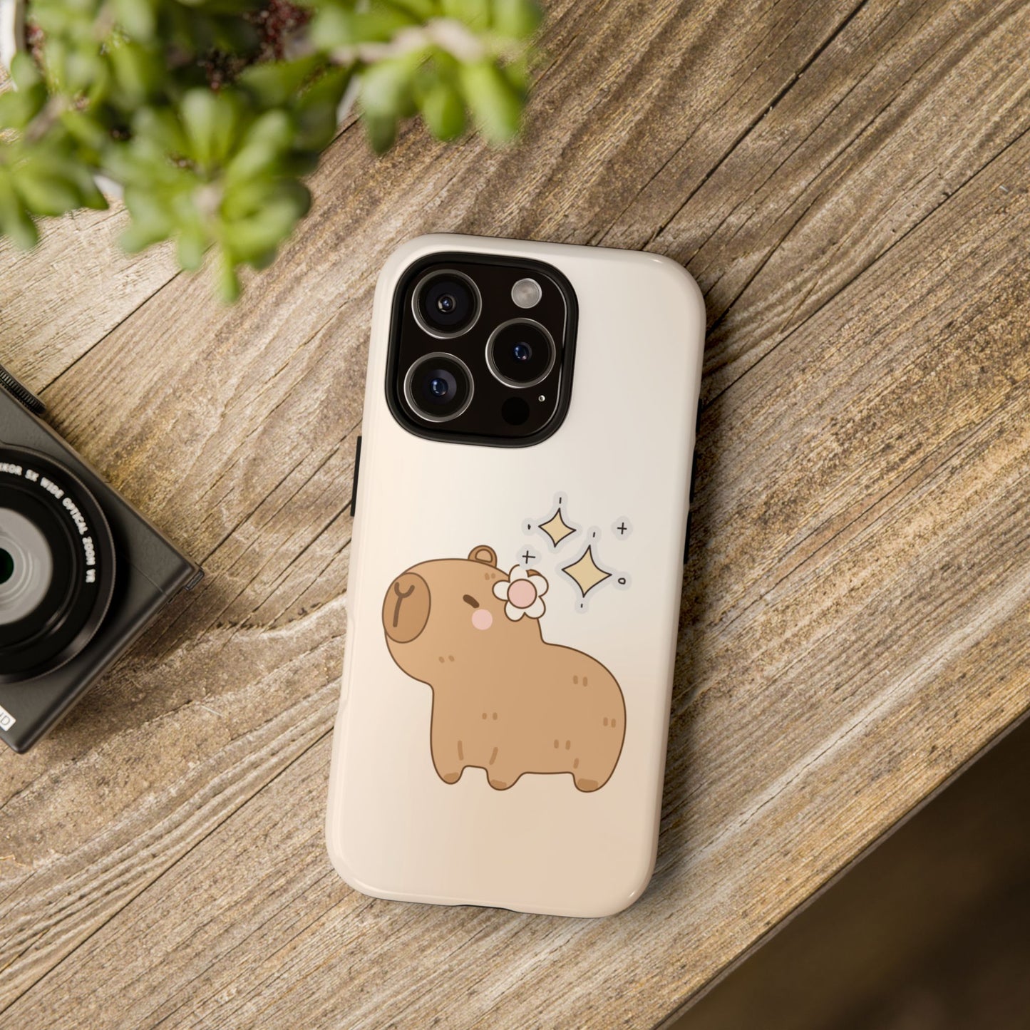 Aesthetic flatlay of kawaii capybara cartoon phone case with flower and sparkles – beige animal-themed cover on wood desk background for iPhone